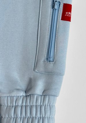 Hellblaue Jogginghose mit einem gerippten Bund, ausgestattet mit einer Reißverschlusstasche in einer passenden Farbe und einem kleinen roten Logotag.