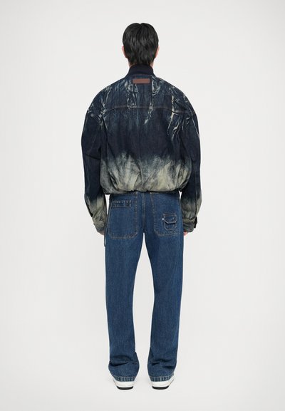 Denim jaka ar tumši indigo un krēmkrāsas gradienta dizainu, saskaņota ar brīva piegriezuma zilajiem džinsiem, abiem demonstrējot teksturētu audumu.