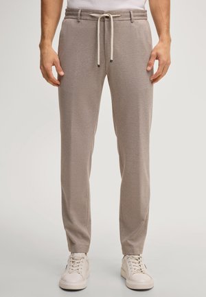 Homme portant un pantalon de jogging marron clair avec cordon et des baskets blanches, debout devant un fond uni.