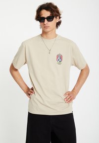 Beige cotton t-shirt with short sleeves, featuring a colorful logo graphic on the chest. Worn with black shorts and sunglasses.