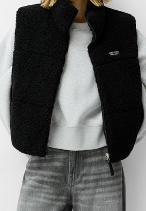 Person iført en sort fleecevest med lynlås over en lys grå sweatshirt og falmede grå jeans mod en ensfarvet hvid baggrund.