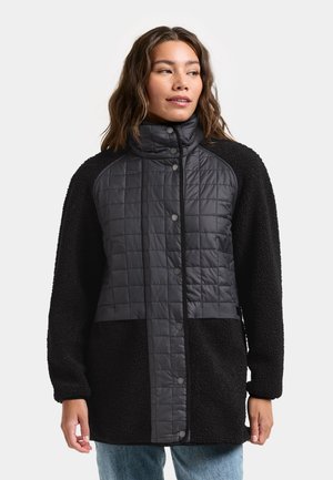 Woman wearing a black quilted and fleece jacket with snap buttons, standing with hands relaxed, looking slightly to the side.