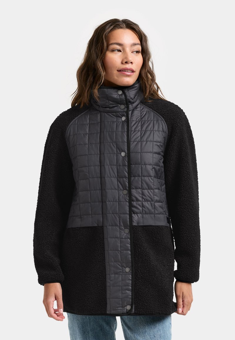 Kvinde iført en sort quiltet og fleecejakke med trykknapper, stående med afslappede hænder, kigger let til siden.