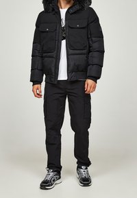 Schwarze Daunenjacke mit Kunstpelzkapuze, ausgestattet mit zwei Fronttaschen, kombiniert mit schwarzen Cargo-Hosen und schwarzen Turnschuhen. Weißes T-Shirt darunter.