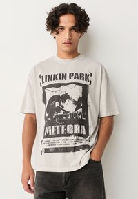 Giovane con capelli ricci che indossa una t-shirt grigia della band Linkin Park Meteora e jeans neri, in piedi con una mano in tasca.