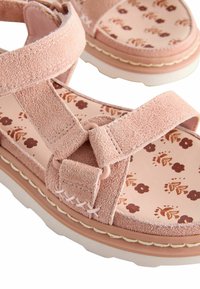 Sandales en suède rose avec des brides croisées, semelle intérieure à motif floral et semelle blanche épaisse avec des détails de couture décoratifs.