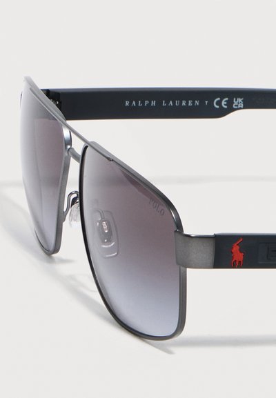 Γυαλιά ηλίου Ralph Lauren με μαύρο μεταλλικό πλαίσιο, φακούς με γκρι διαβάθμιση και κόκκινο λογότυπο παίκτη πόλο στο πλευρικό μπράτσο.