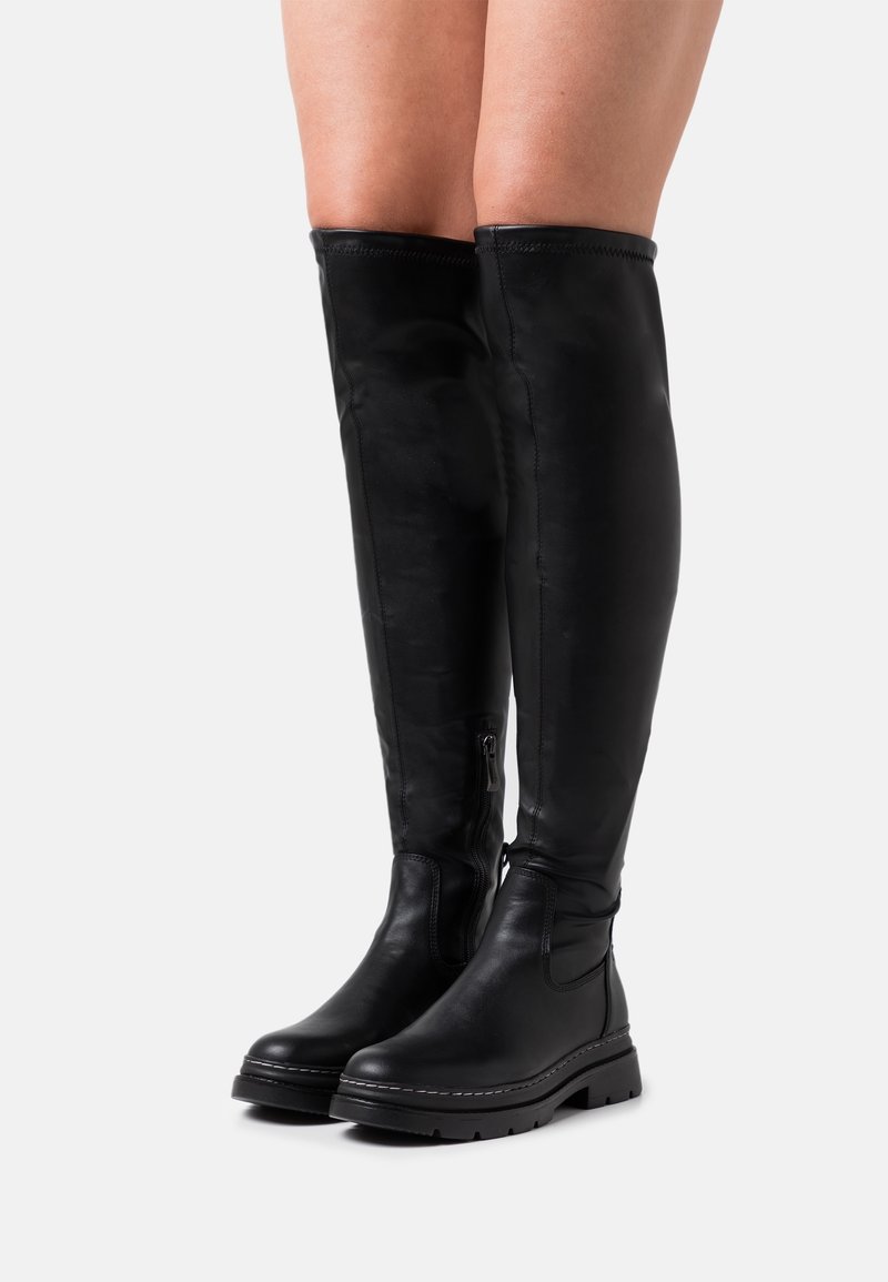 Jambes portant des bottes en cuir noires montantes jusqu'aux genoux, avec des semelles épaisses et des fermetures éclair latérales, posées devant un fond clair uni.
