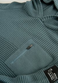 Hoodie bleu clair avec une texture côtière, doté d'une poche poitrine zippée et d'un col structuré. Le tissu semble doux et durable.