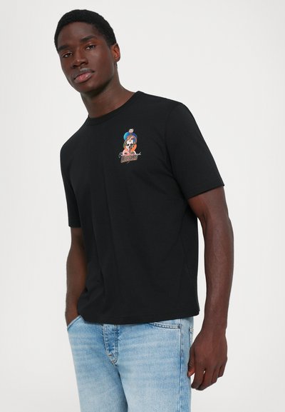 Man wearing a black t-shirt with a cobra and skull graphic on chest, paired with light blue jeans, standing against a plain white background.