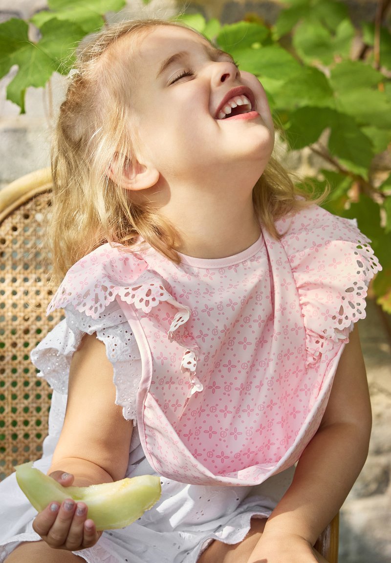 Rosa bib med spetsdetaljer och blommönster, buren av ett barn som håller en bit melon, sittande på en korgstol med grönskande växtlighet i bakgrunden.