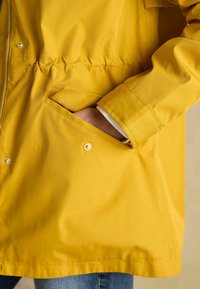 Geltona nepralaidi striukė su tekstūruotu paviršiumi, turinti šoninį kišenę, elastinę juosmenį ir spaudės užsegimus. Matoma prekės ženklo pavadinimas ant apvado.