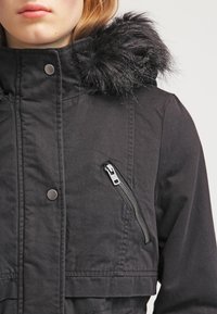 Kobieta w czarnej, ocieplanej kurtce z kapturem wykończonym futrem, z zatrzaskami i ukośną kieszenią na zamek błyskawiczny na piersi, na jednolitym tle.