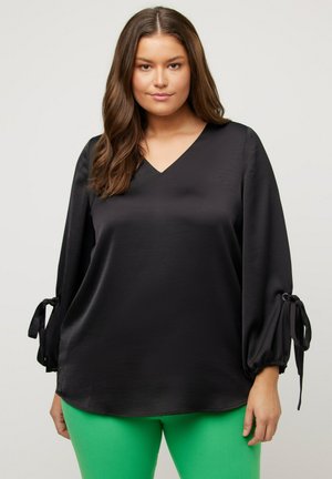 Schwarze Satinbluse mit V-Ausschnitt und Ballonärmeln, ausgestattet mit Satinbändern an den Manschetten; kombiniert mit knallgrünen, körperbetonten Hosen.