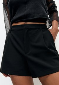 Shorts noirs ajustés avec plis à l'avant, fabriqués en tissu lisse. La taille possède des passants pour ceinture ; un design minimaliste sans motifs supplémentaires.