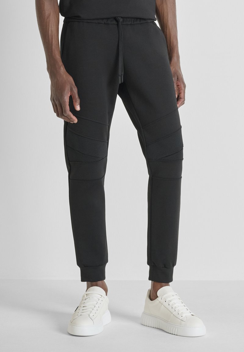 Crne trenirke od mekog materijala, s teksturiranim panelima i suženim krojem. Dizajnirane s elastičnom gumom u struku i manžetama na gležnjevima.