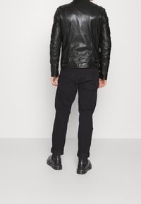 Giacca da moto nera in pelle con maniche con zip, lati con fibbie e una texture liscia e lucida. Indossata con pantaloni neri e stivali alla caviglia.