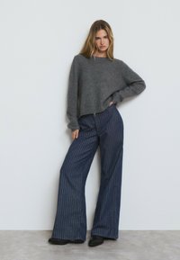 Suéter gris de punto con un ajuste relajado y dobladillo acanalado, combinado con pantalones de mezclilla de pierna ancha a rayas en azul oscuro. Botines negros de tobillo completan el conjunto.