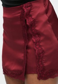 Falda de satén burdeos con una abertura lateral adornada con detalles intrincados de encaje, brillo suave y un dobladillo de encaje con borde festoneado.