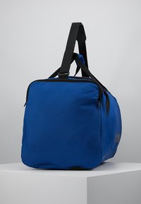 Bolsa de viaje de tela azul con forma rectangular, cremallera y correas negras, que cuenta con un asa superior y una correa para el hombro ajustable.