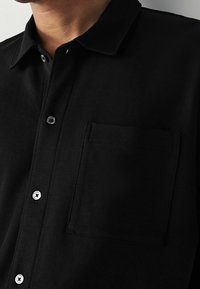 Musta nappikauluspaita, joka on valmistettu pehmeästä kankaasta, sisältää kauluksen, yhden rintataskun ja erottuvat napit visuaaliseksi yksityiskohdaksi.