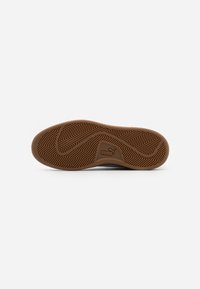 Suola di sneaker in gomma marrone con un motivo a spina di pesce. Presenta una toppa con il logo contenente il nome del marchio in un inserto rettangolare.