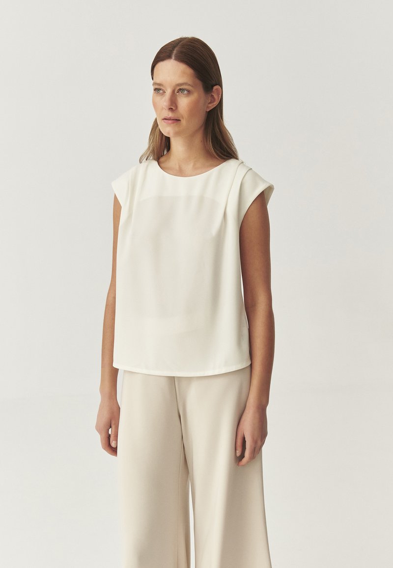 Blusa blanca de mangas cortas hecha de un tejido ligero, con un escote redondo y un corte holgado, combinada con pantalones de pierna ancha en color beige.