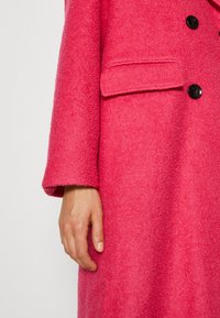 Fuksia vlněný kabát s texturovaným povrchem, který má náprsní kapsu, černé knoflíky a mírně oversized střih.