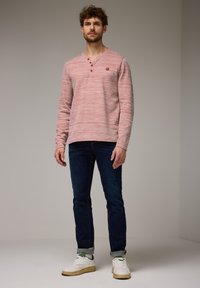 Chemise à manches longues henley rayée rouge et blanche avec patte de boutonnage à trois boutons. Associée à un jean bleu foncé et des baskets blanches. Fond uni.