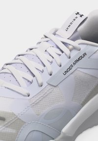 Κοντινή λήψη λευκού παπουτσιού τρεξίματος Under Armour με λευκά κορδόνια, υφή από δίχτυ, και ετικέτα "HOVR" στην γλώσσα του παπουτσιού.