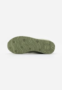 Semelle d'une chaussure vert olive avec des rainures ondulées, des découpes circulaires et le nom de la marque "WODEN" embossé près du talon.