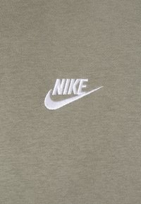 Vit broderad Nike-logotyp med swoosh på en olivgrön stickad tygbakgrund.