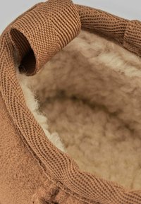 Zapatilla de textil marrón con un interior suave y esponjoso, de forma redondeada y con un acento de tejido trenzado en el borde superior.