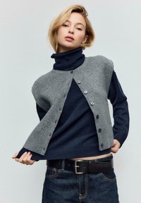 Donna che indossa un maglione dolcevita blu navy sotto un gilet grigio senza maniche con bottoni, jeans scuri e cintura nera, posando su uno sfondo semplice.