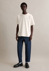 Wit katoenen T-shirt met korte mouwen, met blauwe tekst op de borst, gecombineerd met donkerblauwe spijkerbroek en zwarte schoenen.