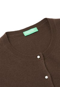 Hnědý pletený kardigan s kulatým výstřihem, který má dva bílé knoflíkové doplňky a zelenou cedulku s nápisem "United Colors of Benetton."