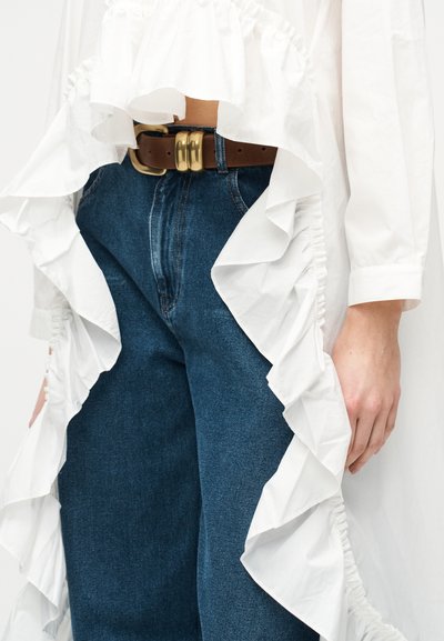 Μπλε τζιν με καφέ ζώνη που διαθέτει διπλή χρυσή πόρπη, φορεμένα με λευκή μπλούζα μακριάς μανίκας με μεγάλα βολάν στις άκρες.