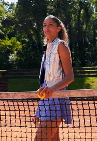 Frau hält einen gelben Tennisball, steht neben dem Tennisnetz auf einem Sandplatz mit Bäumen im Hintergrund, trägt eine silberne Weste und einen blauen Rock.