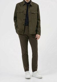 Chaqueta de algodón verde oliva con dos bolsillos en el pecho, cierre de botones, combinada con pantalones marrones oscuros y zapatillas blancas. Ajuste casual.