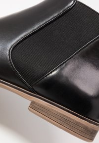 Bota negra con parte superior de cuero liso, paneles laterales elásticos y un tacón de madera apilada con un sutil patrón de vetas.
