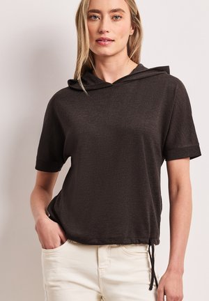 Blonde vrouw draagt een zwart hoodietje met korte mouwen en een trekkoord in de zoom, en lichtbeige broek, staand met één hand in de zak.