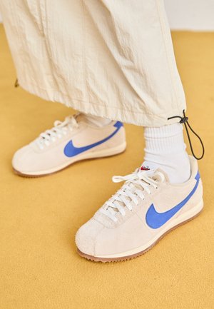 Beige sneakers med blå Nike-swooshes båret med hvide sokker og løse cremefarvede bukser på en gul gulv.