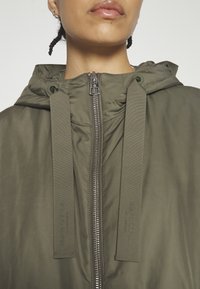 Gros plan sur une personne portant une veste à capuche vert olive avec une fermeture éclair frontale et de larges cordons de serrage.