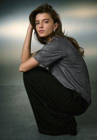 Čierne nohavice so strieborným bodkovaným vzorom a metalicky sivé tričko s krátkym rukávom. Osoba sedí a predvádza jemný lesk a textúru.