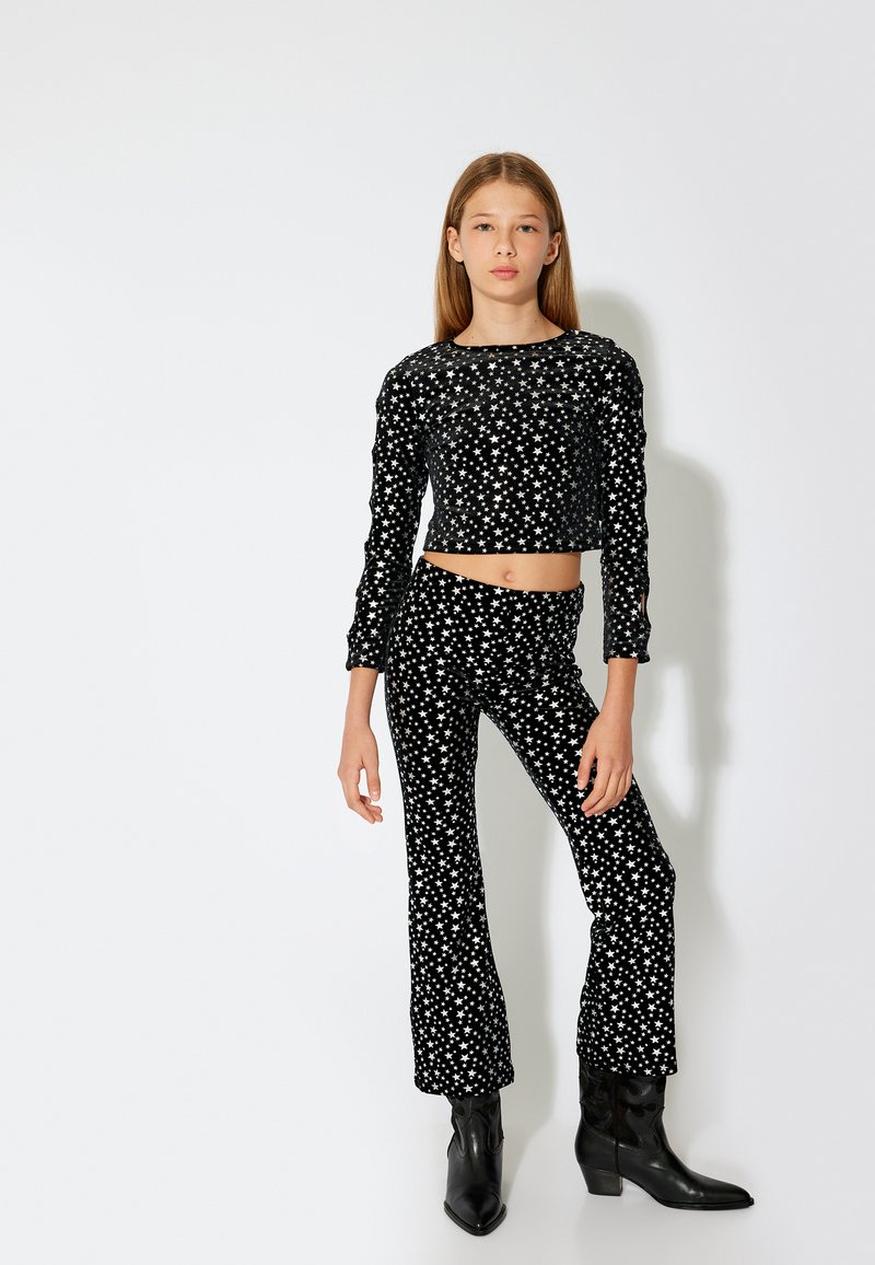 Sort velour to-delt outfit med en cropped langærmet top og flared bukser, prydet med hvide stjerne-mønstre. Sorte hæl støvler fuldender looket.