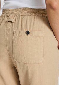 Pantalones de algodón beige con cintura elástica, bolsillos laterales y un bolsillo trasero con parche y un acento de botón oscuro. La textura parece suave.