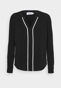 Blusa nera a maniche lunghe con scollatura a V, dettagli in piping bianco lungo la parte anteriore e pieghe strutturate sulle spalle. Tessuto morbido.