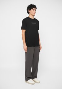 Uomo in piedi che indossa una T-shirt nera di Armani Exchange, pantaloni grigi larghi e sneakers bianche su uno sfondo bianco uniforme.