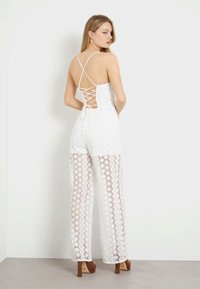 Balta nėriniuota šokinėjimo suknelė su sukryžiuota nugaros dalimi, plačiomis kojomis ir gėlėmis abipus, derinama su rudi aukštakulniai.