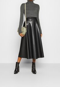 Gonna midi nera in pelle, vita aderente, finitura opaca, abbinata a un top grigio scuro a maniche lunghe e stivaletti neri con tacco, mentre tiene una borsa verde.